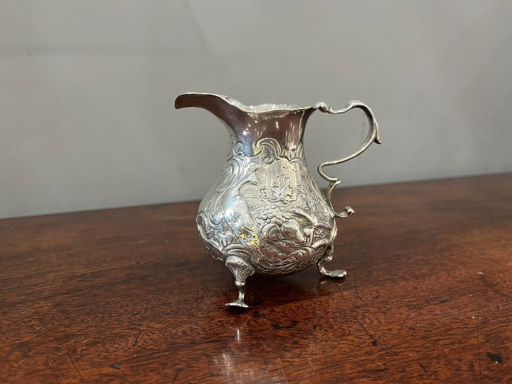 Cream jug London 1755. Shaped rim and everted lip above a repousse decorated body with a C scroll form handle; the whole supported on three trifle feet. (BN793)